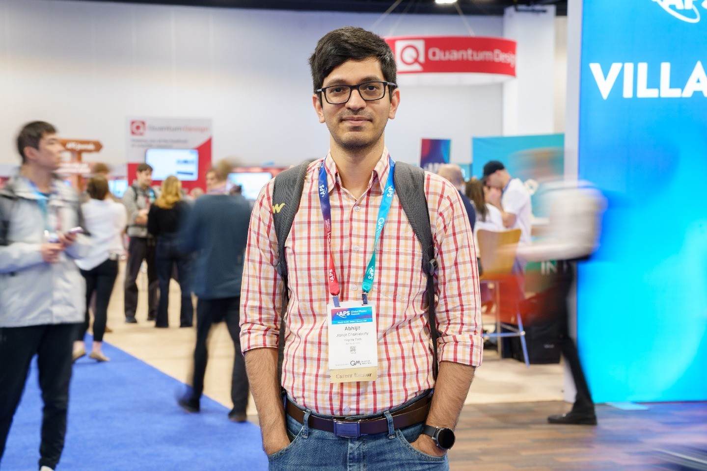 Chakraborty, wearing a colorful lanyard at the APS Global Physics Summit, stands in the summit’s exhibit hall, with moving people appearing as blurs behind him.