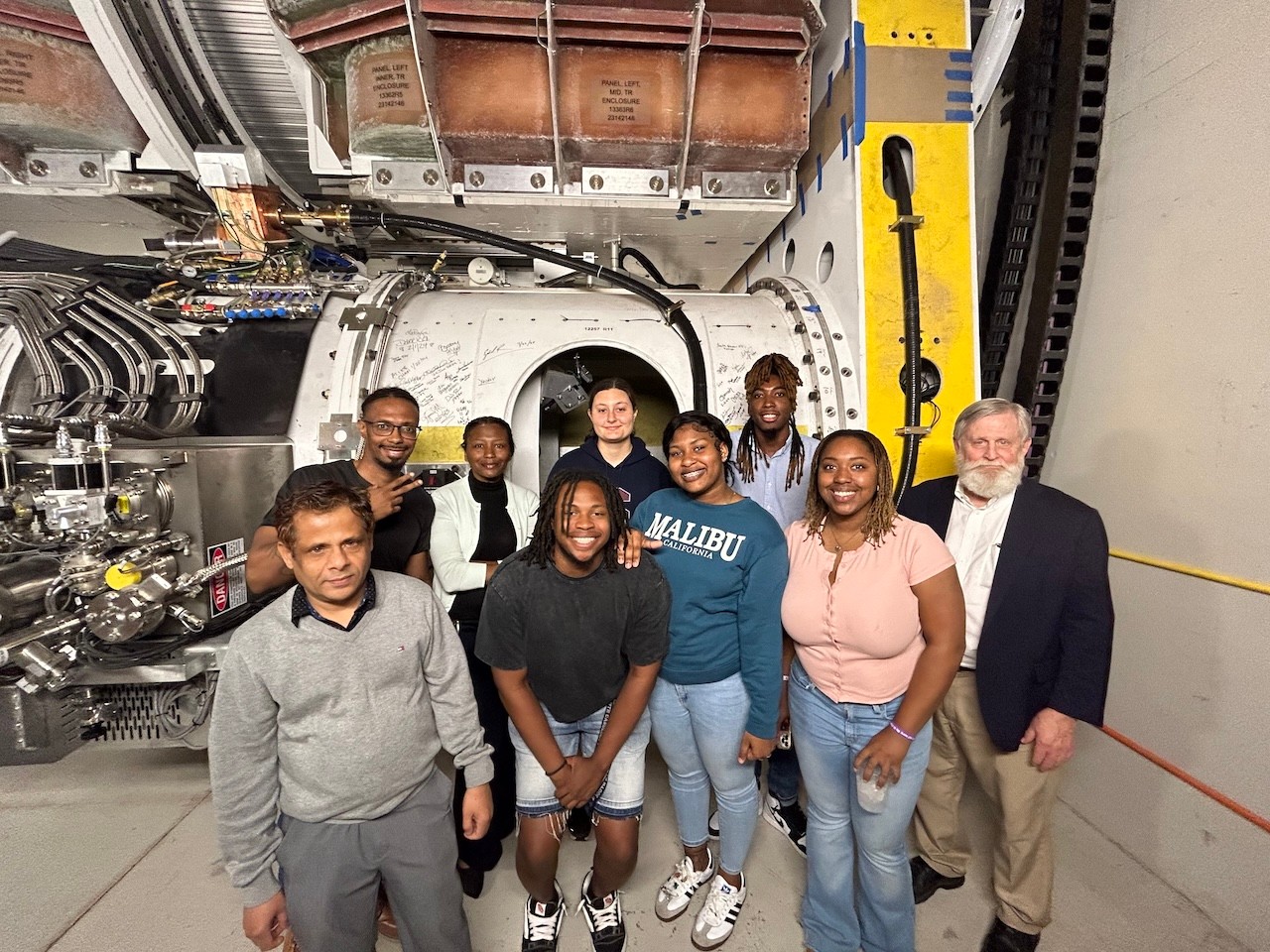 A group of physicists – two professors and seven students – pose in front of a large, cylindrical piece of research equipment, encircled by tubes and wiring.