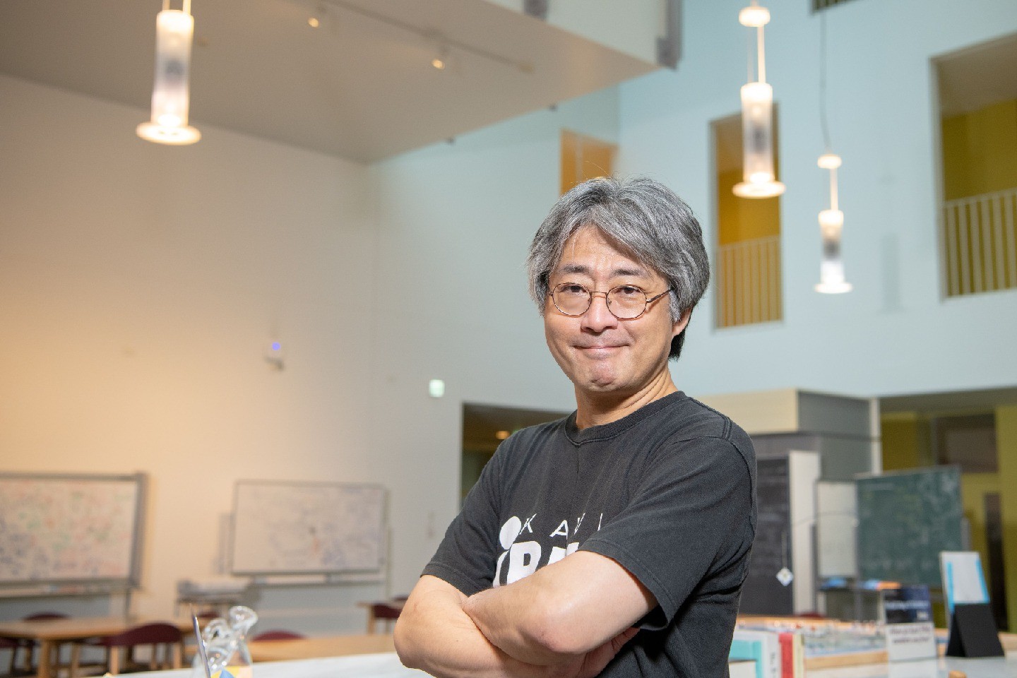 Murayama stands with his arms crossed in a brightly lit room with a high ceiling.