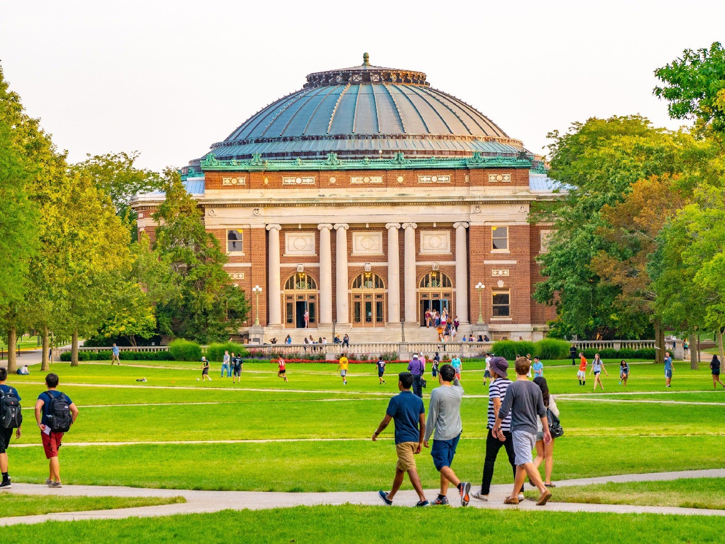 Students walk across a green lawn on a college campus, with a historic, dome-topped building in the background.