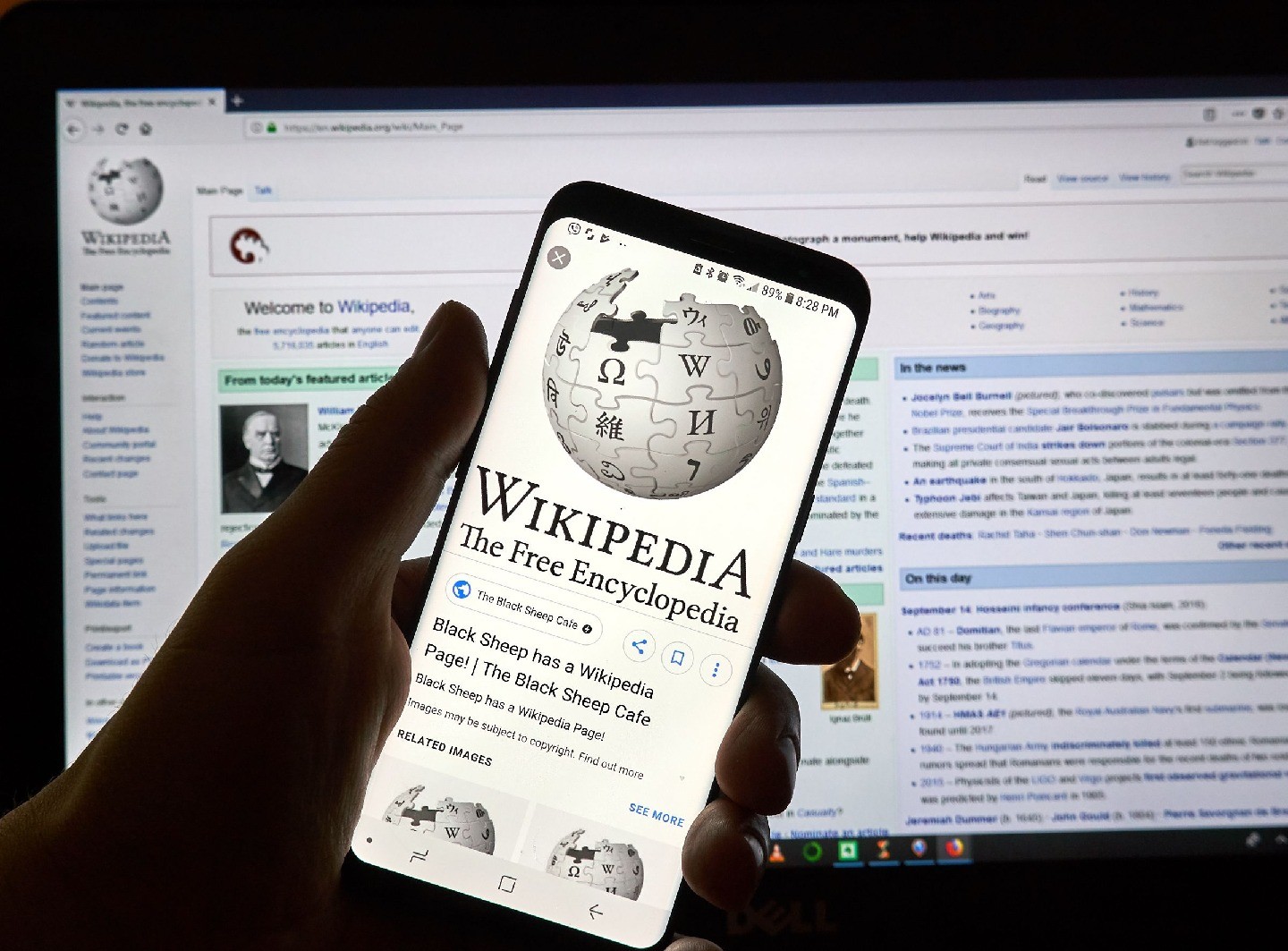 Someone’s hand holding a phone with a Wikipedia page visible on it. Behind the hand, a computer screen is visible, also with a Wikipedia on it.