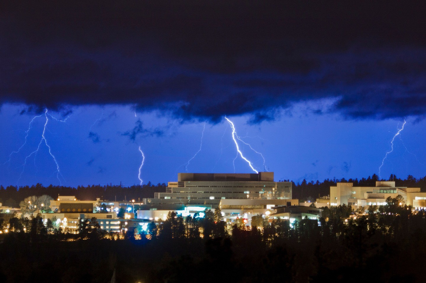 The sprawling Los Alamos Laboratory building is brightly lit under a cloudy, dark blue sky at dusk. Flashes of lightning are visible in the background.