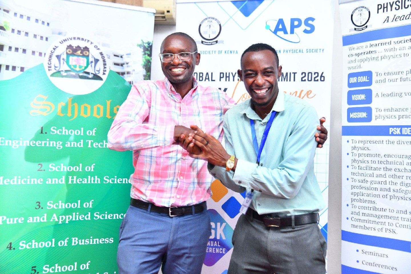 Two summit attendees shake hands and smile in front of APS satellite site signage.