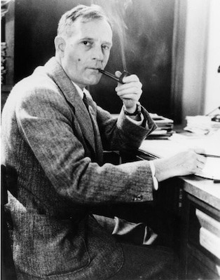 A man seated at a desk, smoking a pipe and posting for a photo.