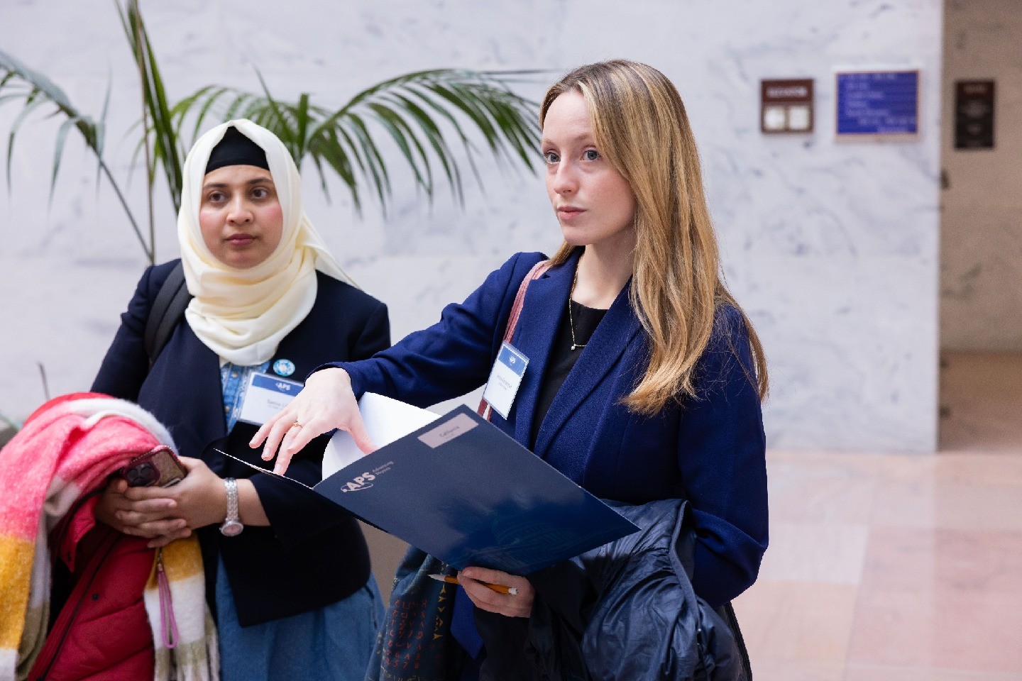 Two attendees stand in a congressional building, one with an APS-branded folder.