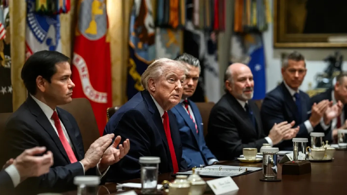 Trump sits at a long table between several Cabinet members, who are applauding.