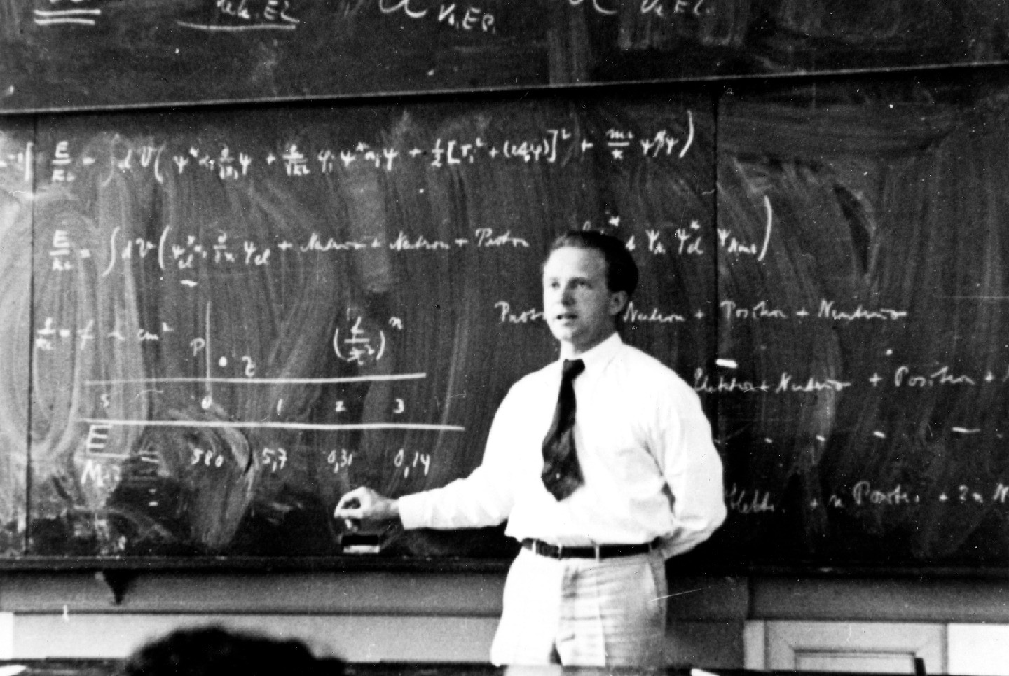 Black-and-white photo of a man holding a piece of chalk and standing with his back to a chalkboard of equations.
