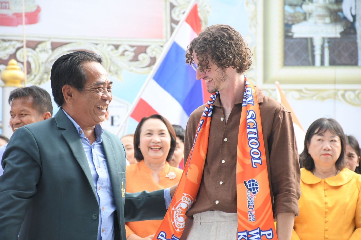 Jack Stillman, with an orange sash around his neck that reads “PWK,” smiles and greets another person at the school in Thailand.