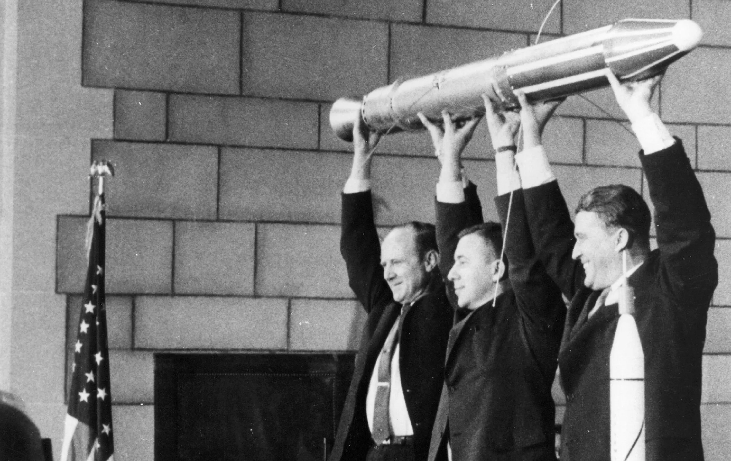 A black-and-white photo shows the three men in suits behind a desk, holding above their heads a replica of the cylindrical Explorer 1. A crowd looks on.
