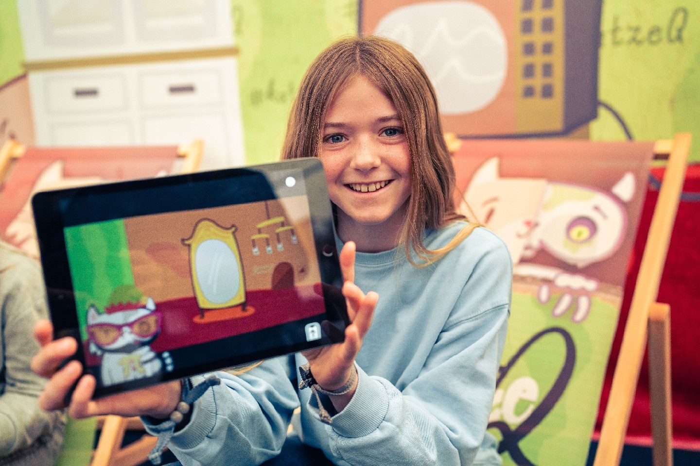 A young smiling girl holds up an iPad with kitty Q visible on it.