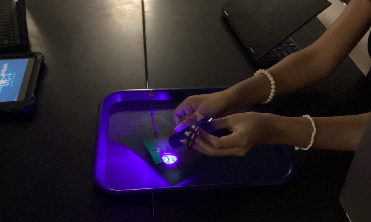 A student – whose hands are visible – shines a flashlight onto a material on a tray, casting a purple light.