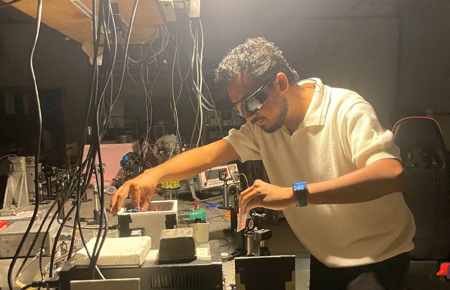 Vithanage, wearing safety glasses, leans over and manipulates specialized equipment in a lab, with cords dangling above him.