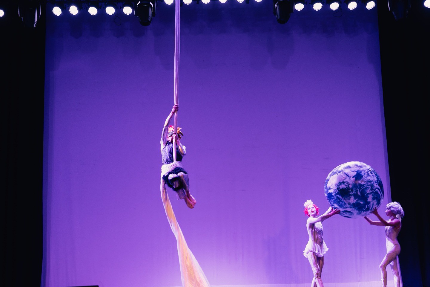 An aerial silk performer is suspended high above stage while two dancers below hold a large ball resembling Earth against a purple backdrop.