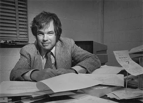 Black and white portrait of Kenneth Wilson sitting at a desk covered with books and papers. Wilson smiles gently at the camera and is wearing a tweed suit with a tie. 