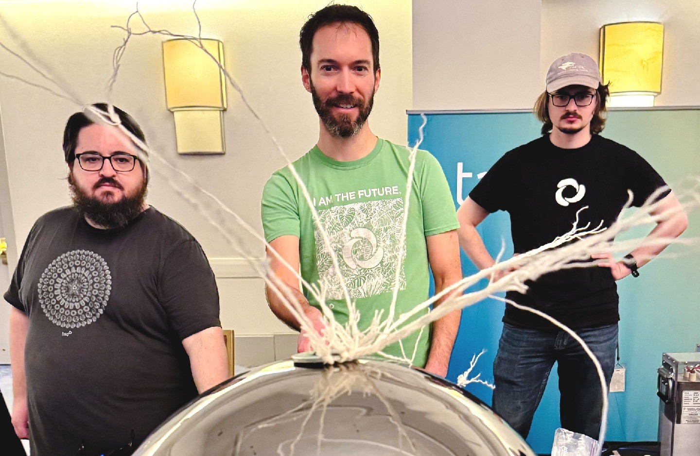 Three people stand behind a shiny, reflective metal sphere from which stringy white fluid branches upward.