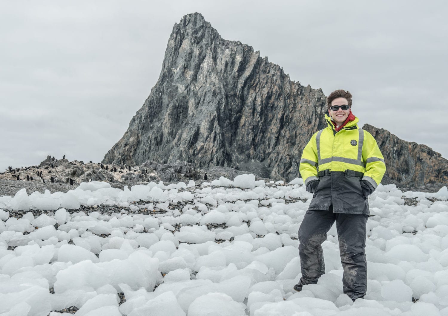 Heather Lynch smiles at the camera. She is wearing sunglasses and a heavy, neon-yellow and black coat and thick black snow pants. She is standing in the middle of a field of scattered pieces of ice. In the background, a rocky stone cliff rises abruptly out of the ground behind a group of dozens of penguins.