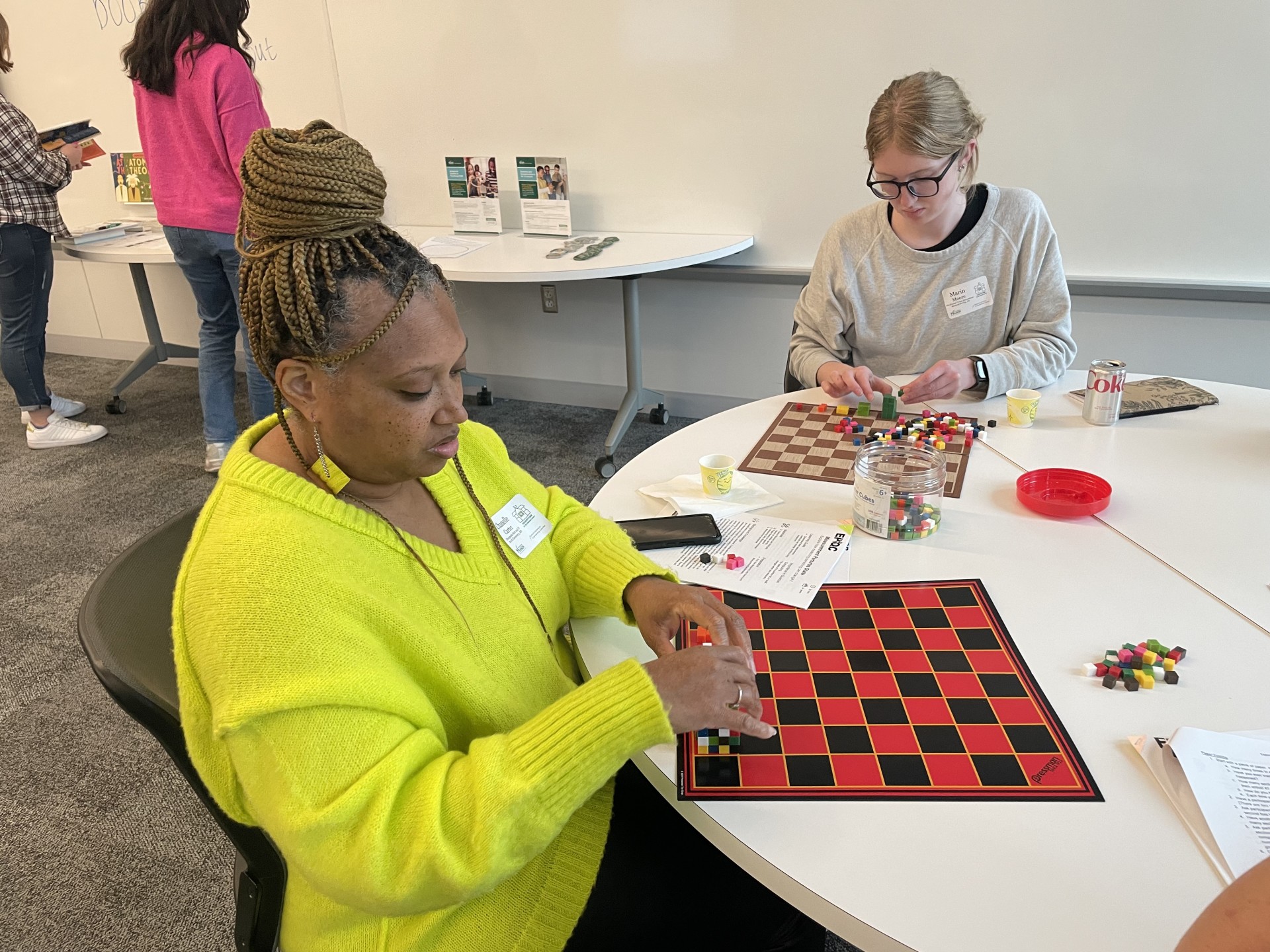 Two teachers are seated at a round table, each focused on their own tabletop activity. Each teacher arranges small, variably colored cubes on a square checkerboard.