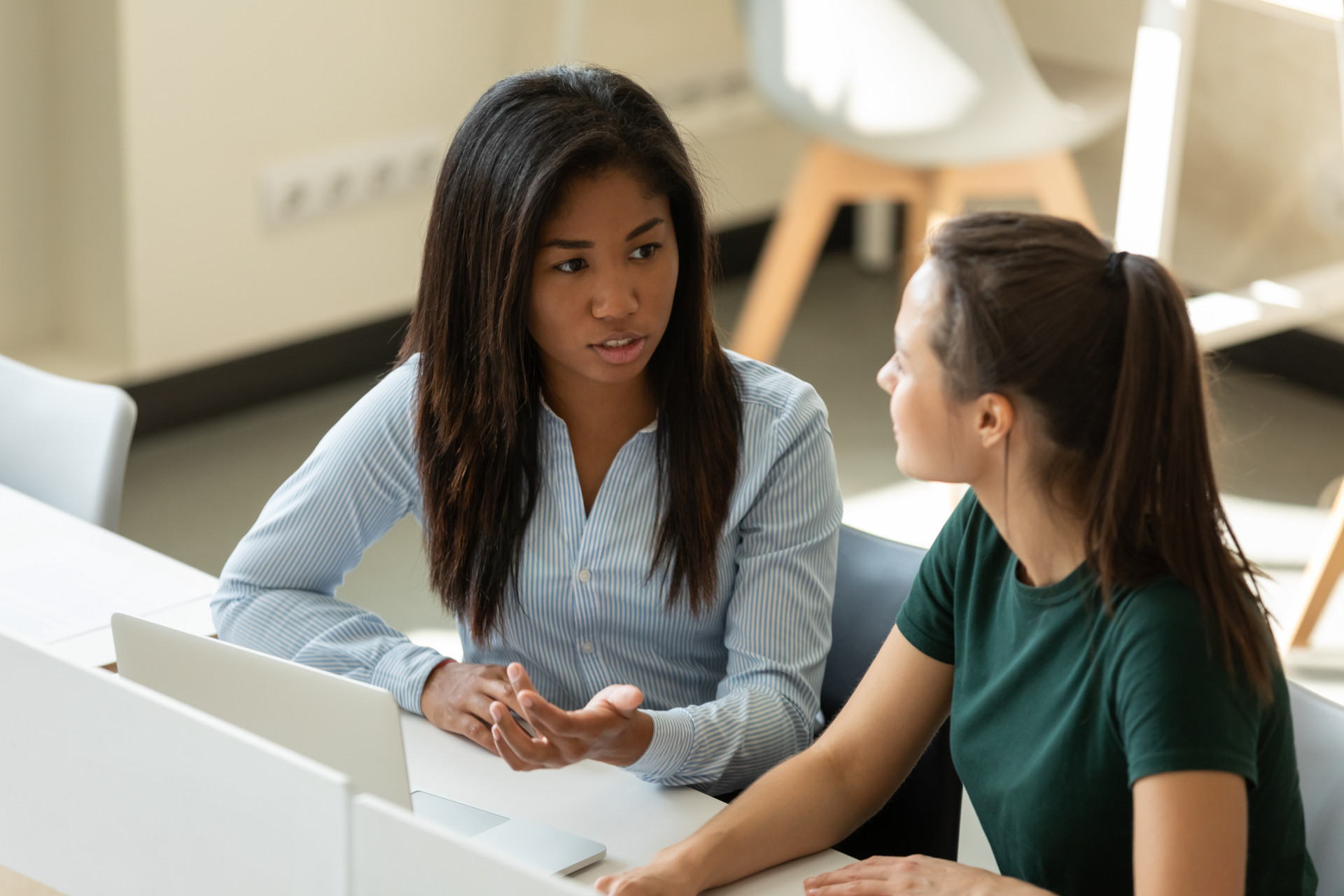 Two women sit at a desk talking