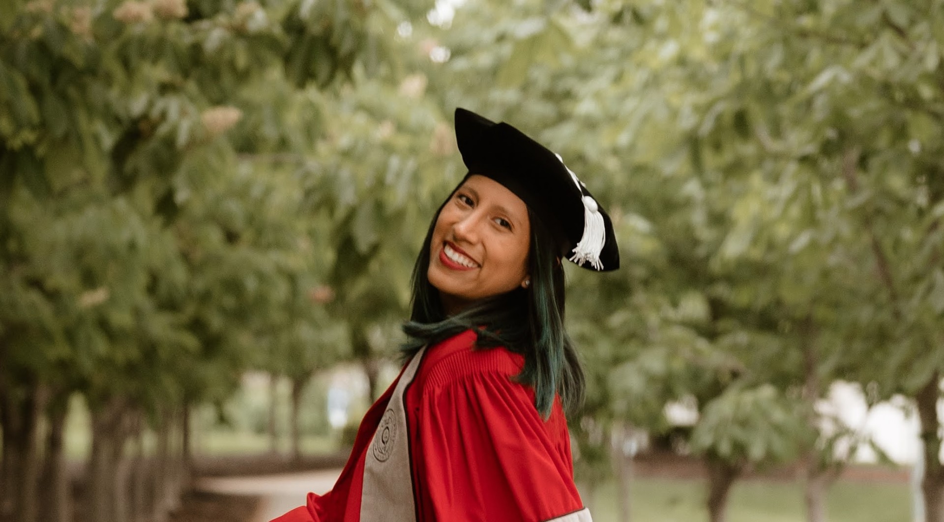 Daniella Roberts looks over her shoulder, smiling at the camera, with trees in the background. She’s wearing a graduation cap and red graduation gown.