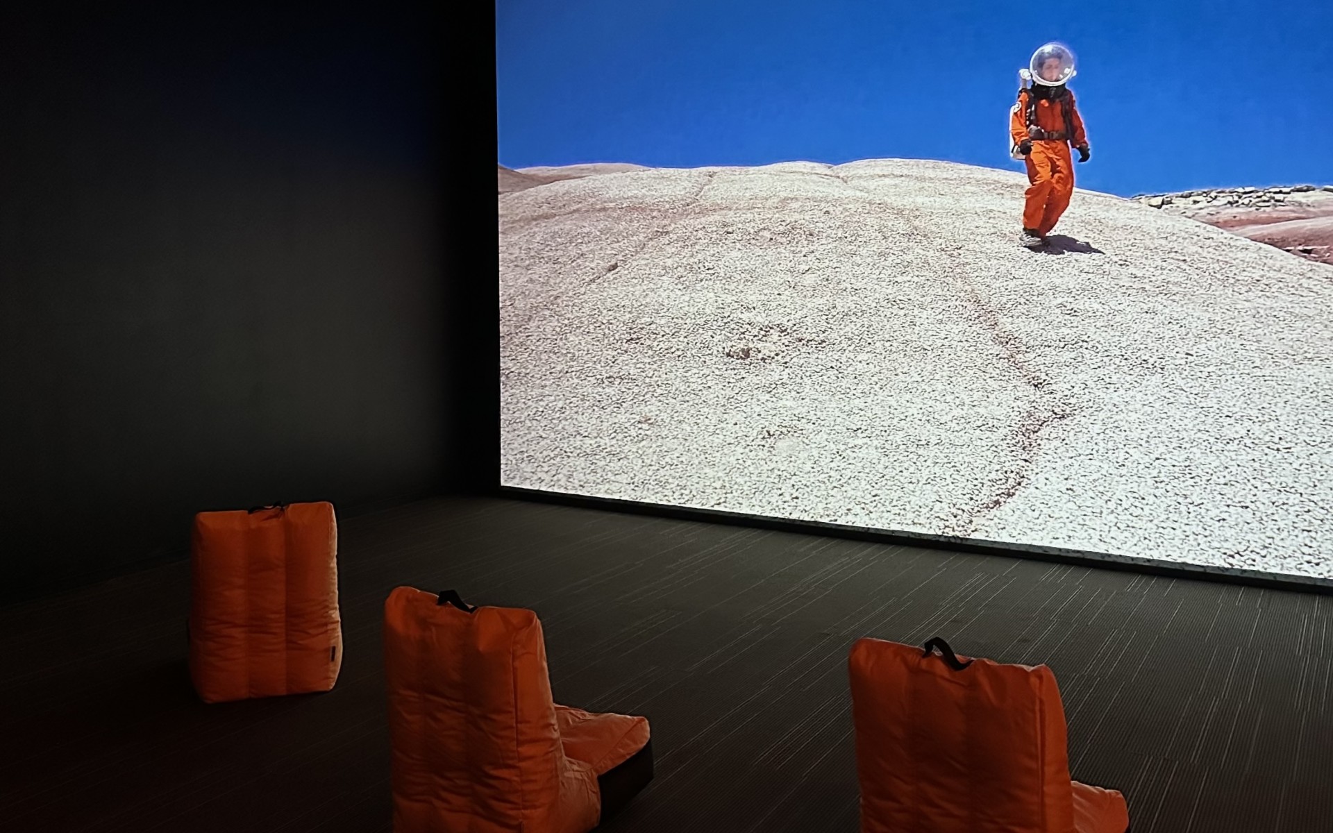 Three orange chairs in front of a large screen