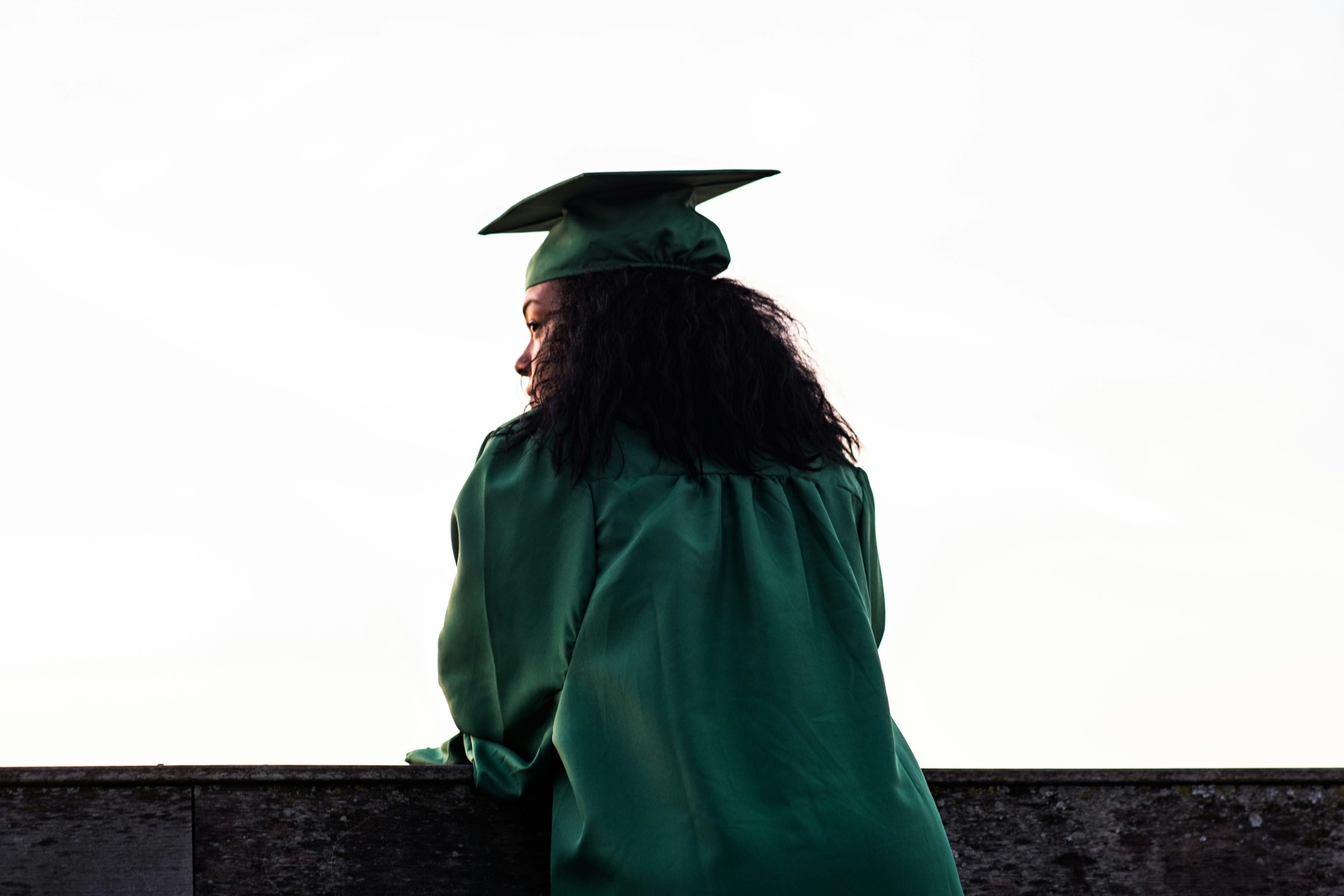 A woman, viewed from behind, is wearing a green graduation gown and cap. She leans on a wall and peers out pensively.