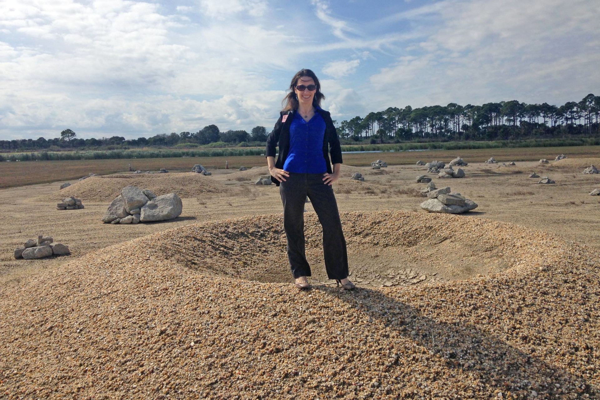 Laura Forczyk, wearing sunglasses on a sunny day and smiling at the camera, stands on the raised edge of a sandy impact crater. Trees are visible in the distance.