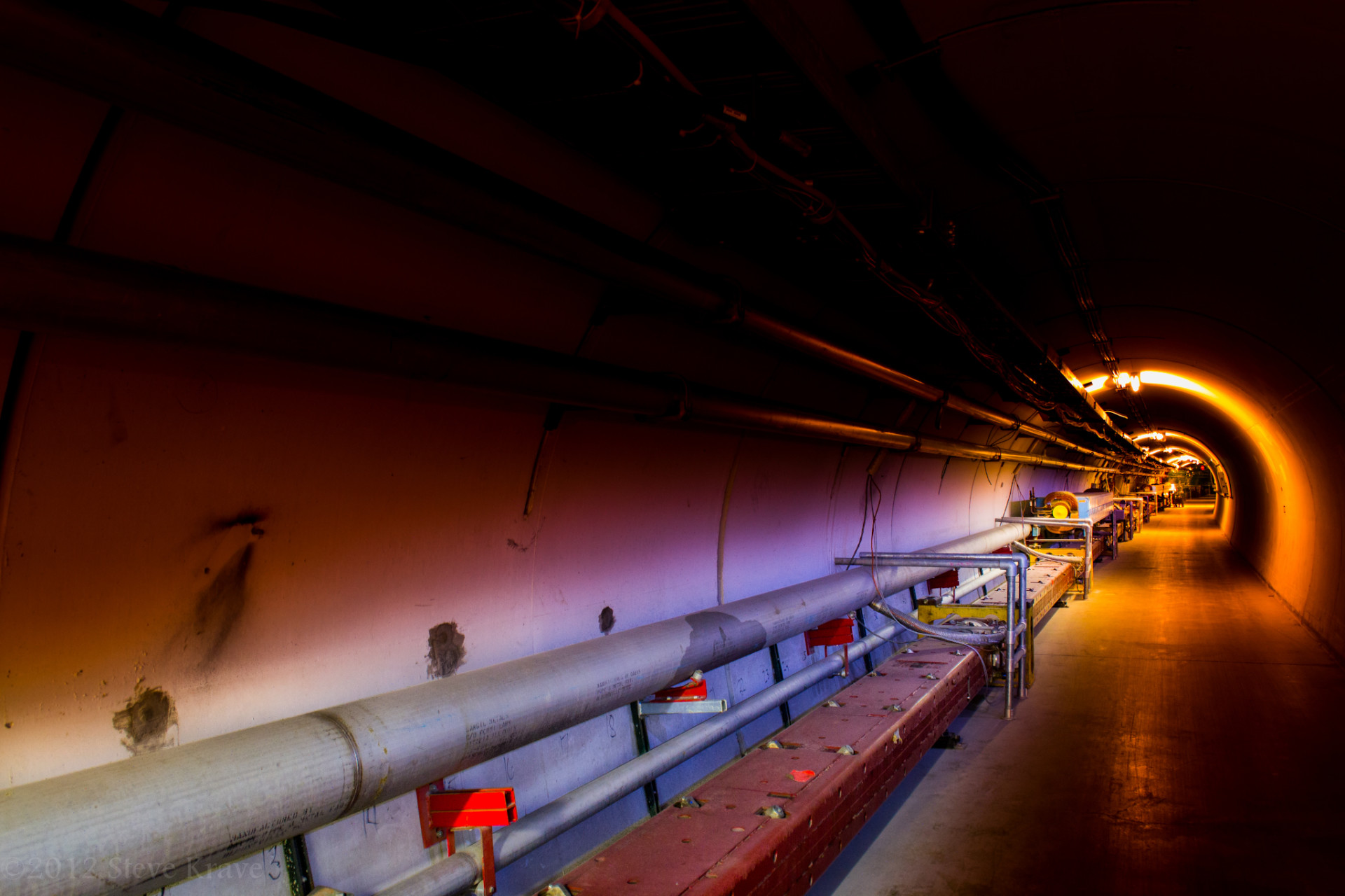 A curved underground tunnel with pipes along the wall and red equipment near the floor, lit by orange lights that fade into darkness.