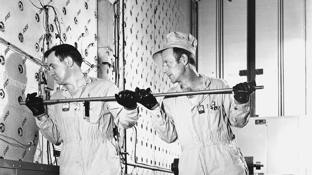 Black-and-white photo of two workers in white coveralls using a long rod to load uranium fuel into a wall that appears covered with a grid pattern of holes. The men are working side by side, gripping the rod together.