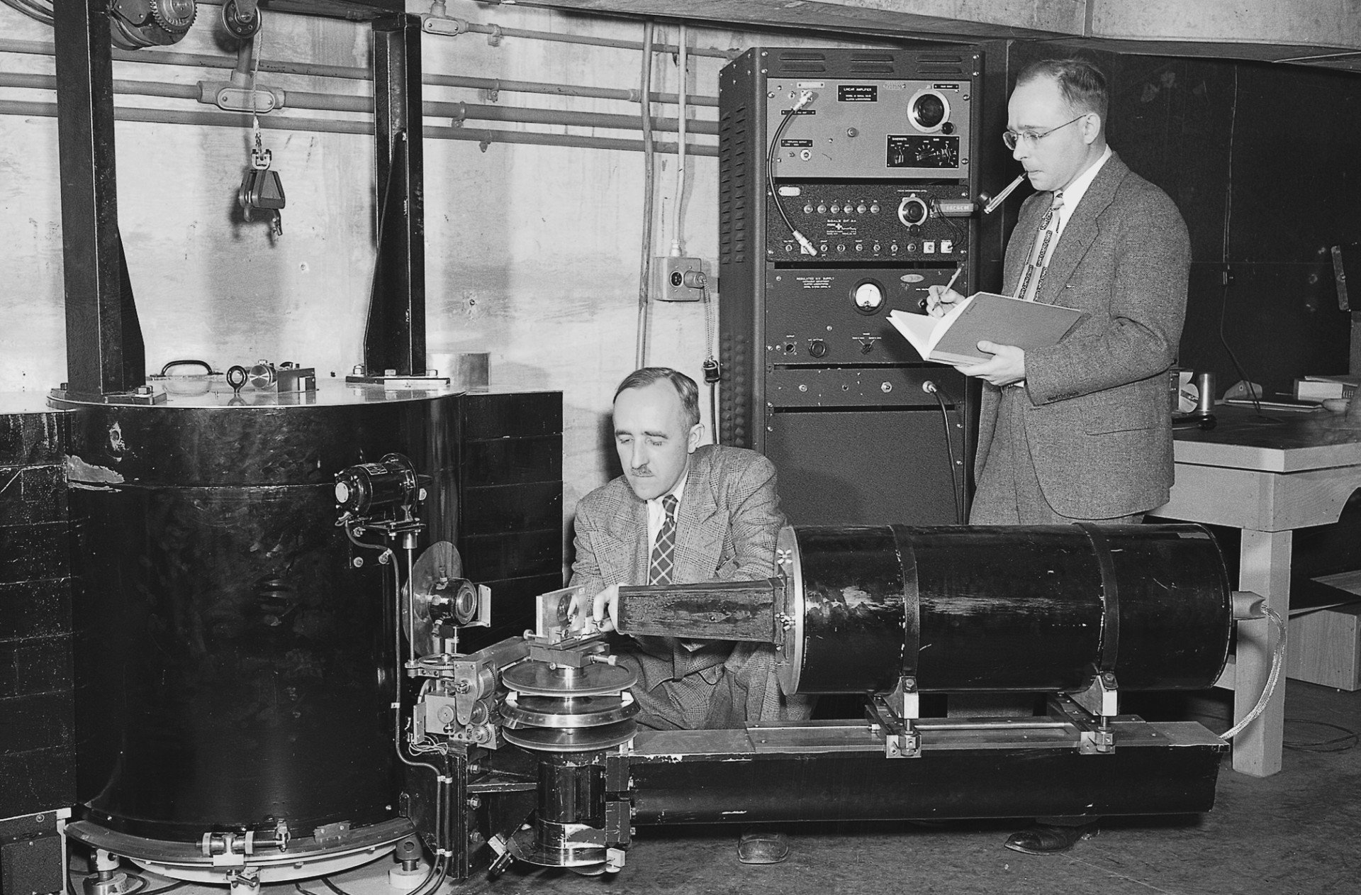 Black-and-white photo of two scientists operating a large, round, metallic-looking neutron diffractometer in a laboratory. Wollan crouches and works on a piece of equipment, while Shull stands taking notes beside large, boxy electronics equipment.