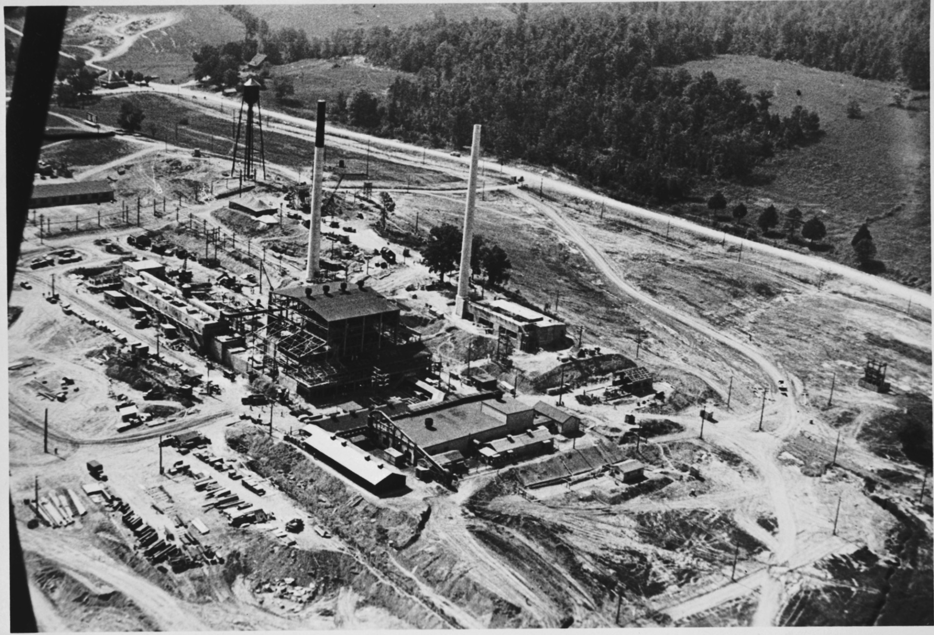 Black-and-white aerial photograph of an industrial facility with two tall smokestacks, surrounded by cleared land and forests. The site includes multiple buildings, access roads, and parked vehicles in a rural setting.