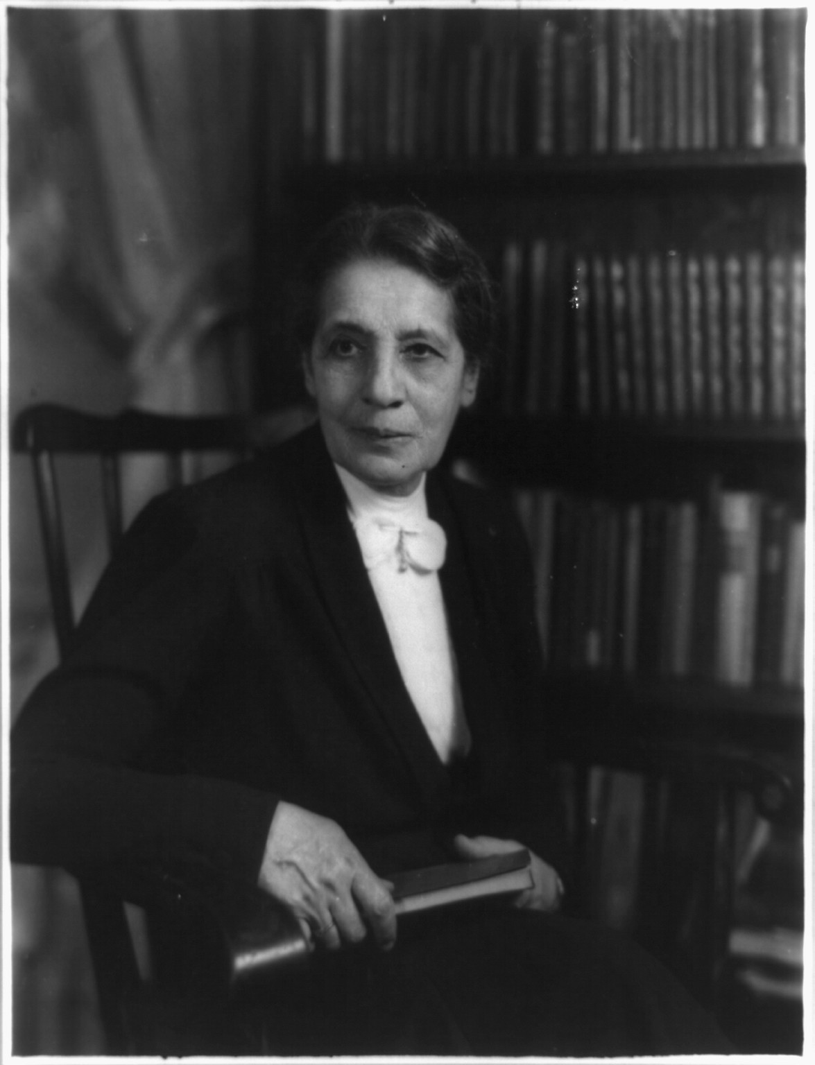 A black-and-white portrait of Lise Meitner seated in a chair with a book in her hand, a full bookshelf in the background.