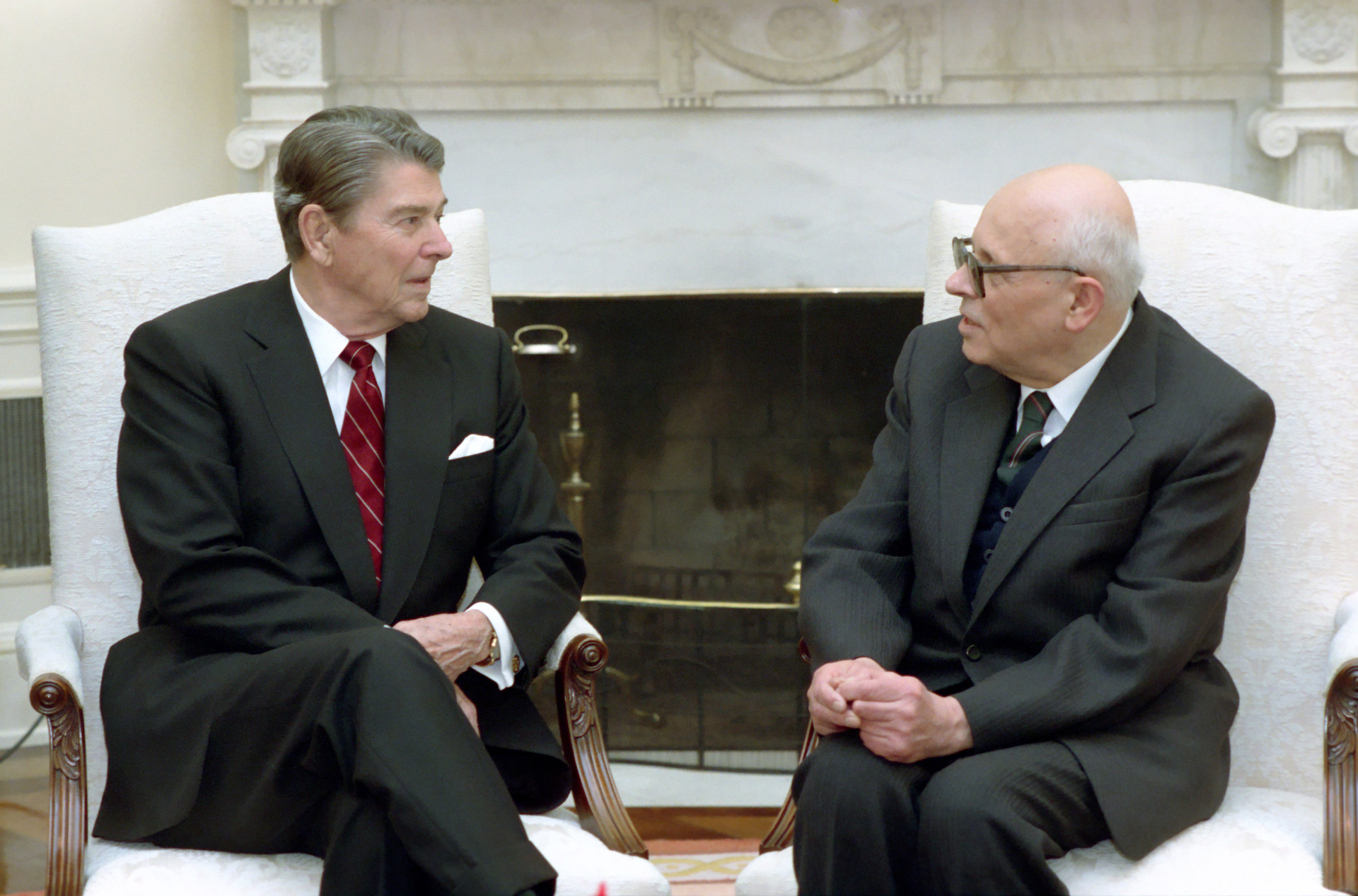Reagan and Sakharov converse. The men are wearing dark suits and seated in white chairs by a fireplace.