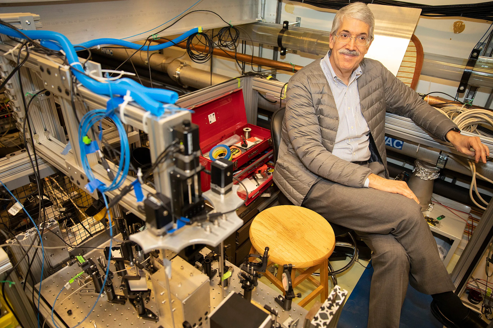 Doyle smiles from a chair in his lab. Complex equipment with blue cables and metal components fills the foreground, with a wooden stool beside him.