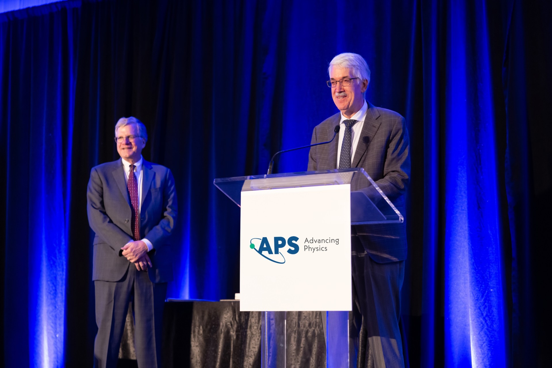 Doyle speaks at a podium with the APS logo. Jon Bagger, the CEO, stands in the back left. Blue stage lighting illuminates the background.