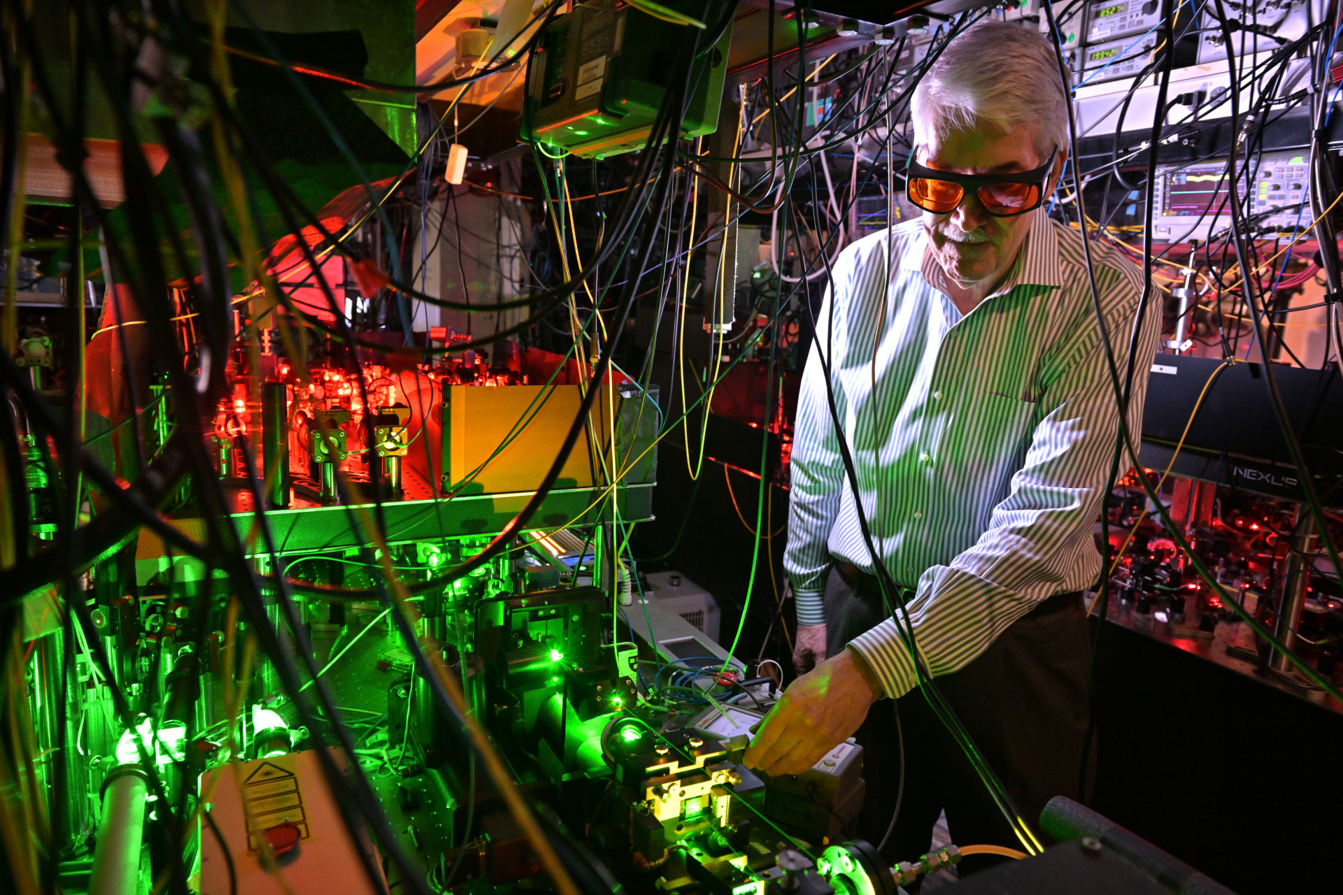 Doyle, wearing red safety glasses, examines a complex laser setup in a dark lab. Green and red lights illuminate equipment in front of him, while cables and wires crisscross the foreground and background.