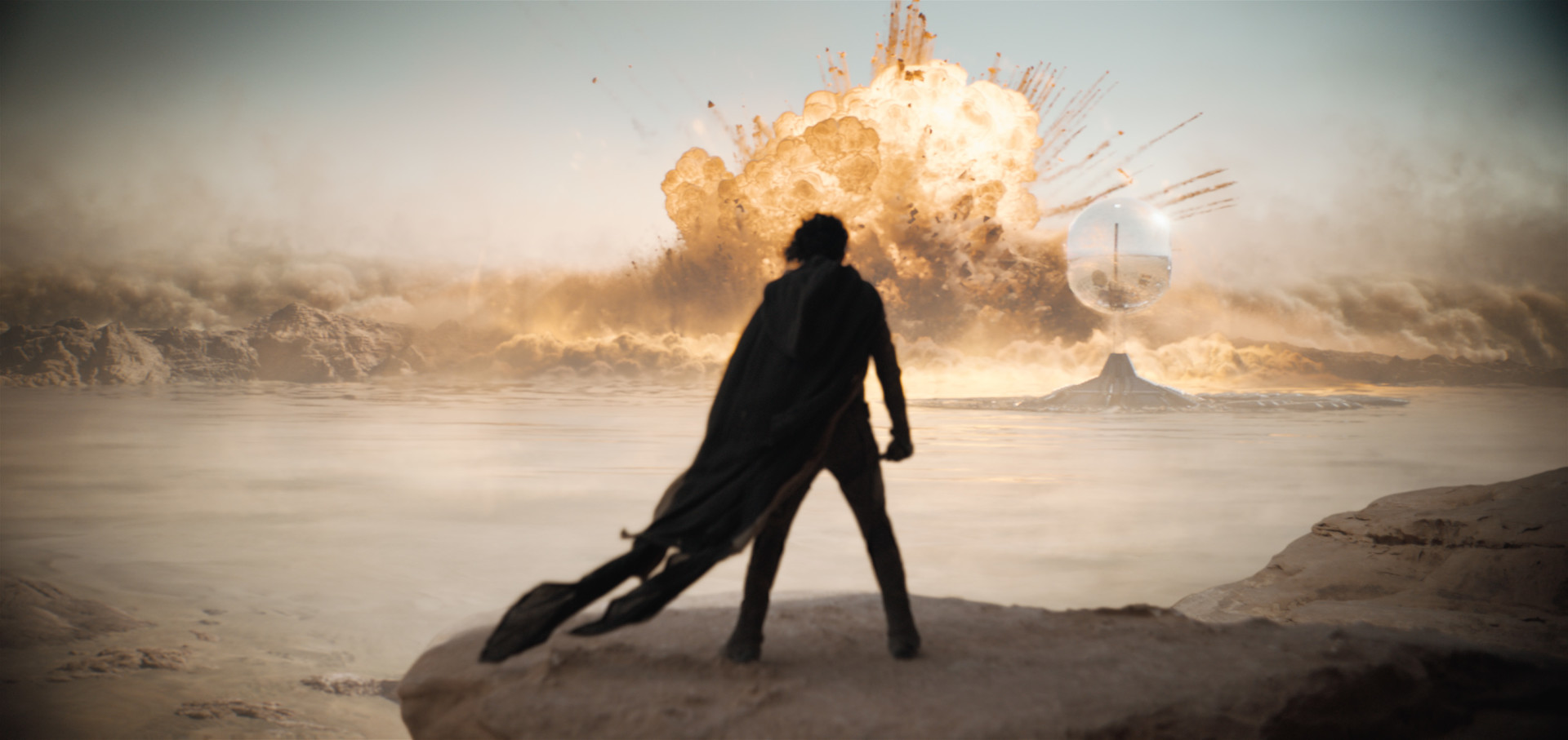 A scene from the film showing the silhouette of character Paul Atreides with flowing cape standing on rocky terrain, watching a massive explosion in a desert landscape with dust clouds billowing in the distance.