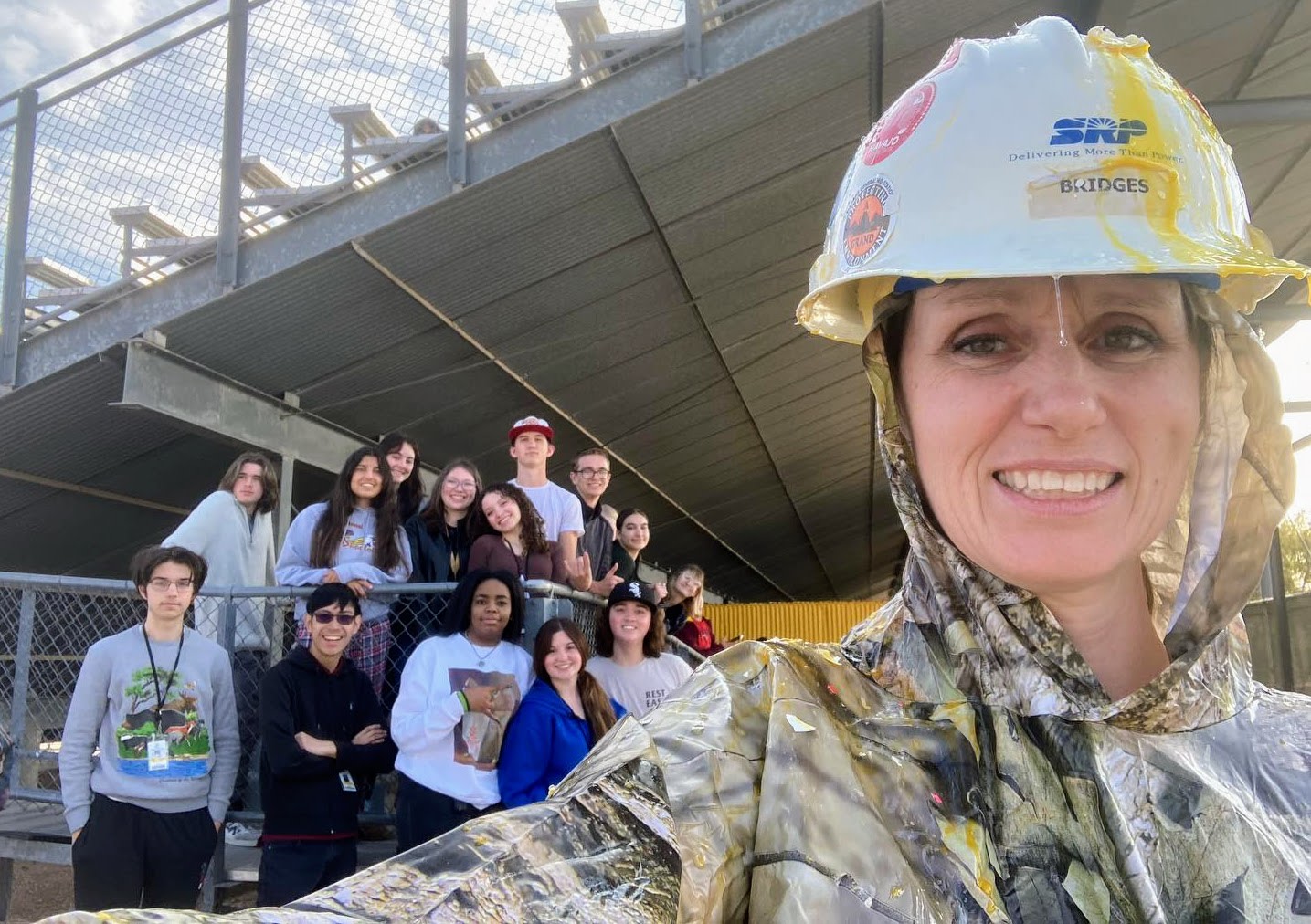 Lynn Jorgensen, wearing a camo poncho and a hard hat with raw egg running dripping off, smiles at the camera with a group of students in the background.