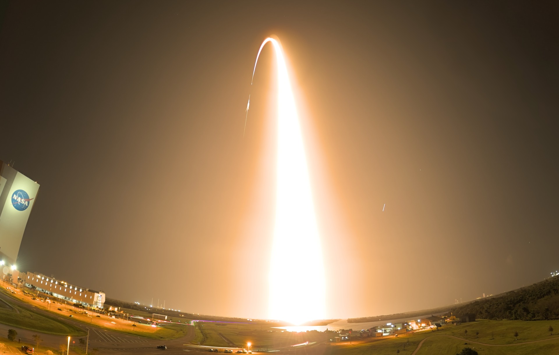A rocket launch at night, with the bright curved trajectory of the rocket visible against the dark sky. NASA logo visible on a building to the left.