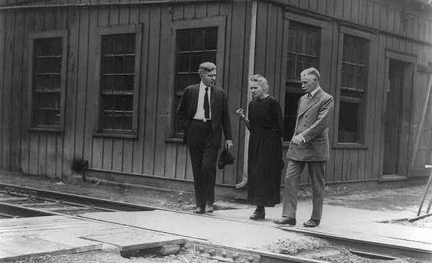 A black-and-white photo of Marie Curie flanked on either side by two men in suits. The trio is conversing and walking across a train track with a building in the background.