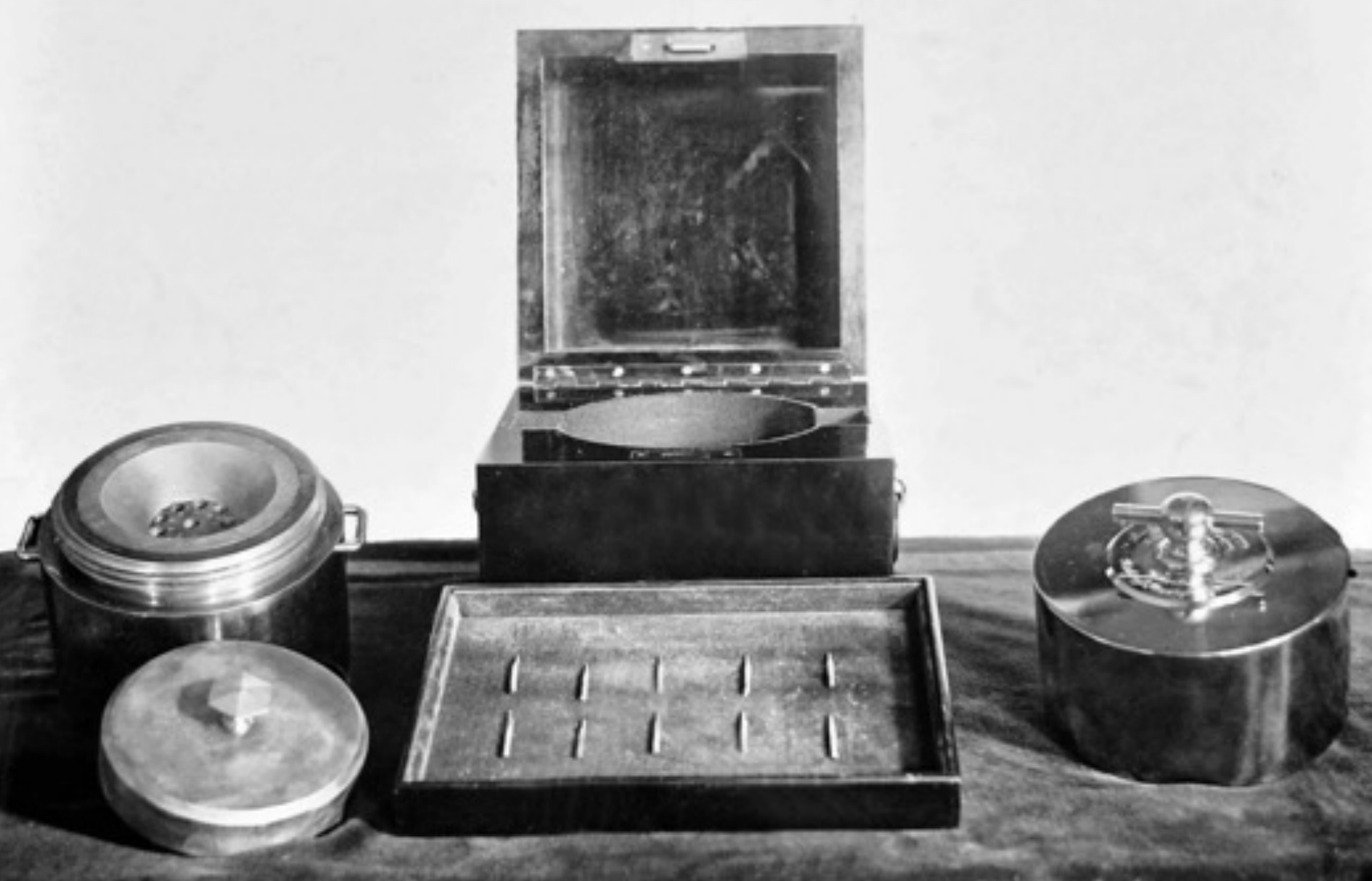 A black-and-white photo shows a neat arrangement of 10 small vials in a shallow rectangular box. Parts of a cylindrical chronometer sit at left and right, and a thick box sits open in the background.