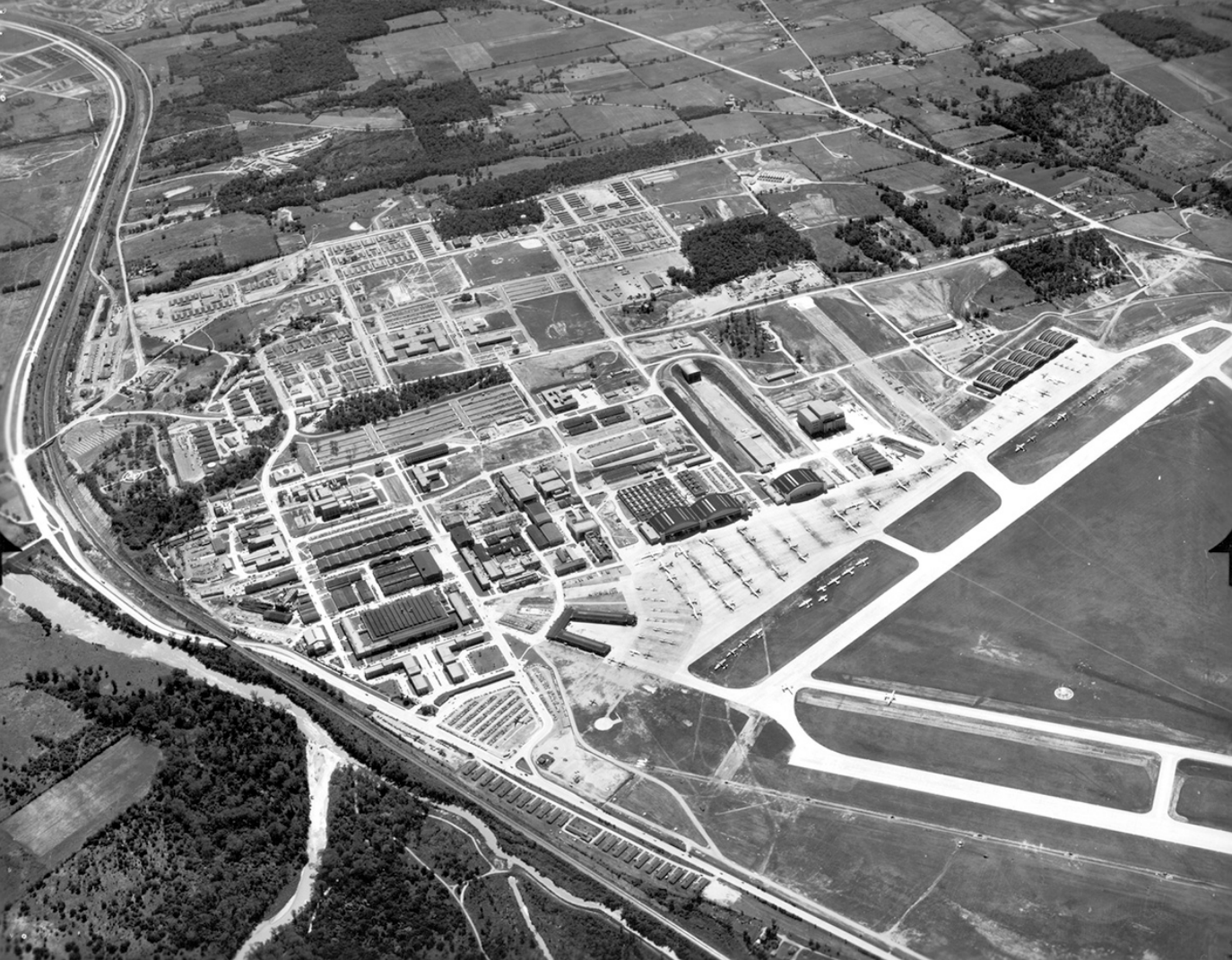 A black-and-white photo shows an aerial view of a sprawling air force campus with buildings, roads, runways, and planes, all surrounded by a patchwork of fields and woods.