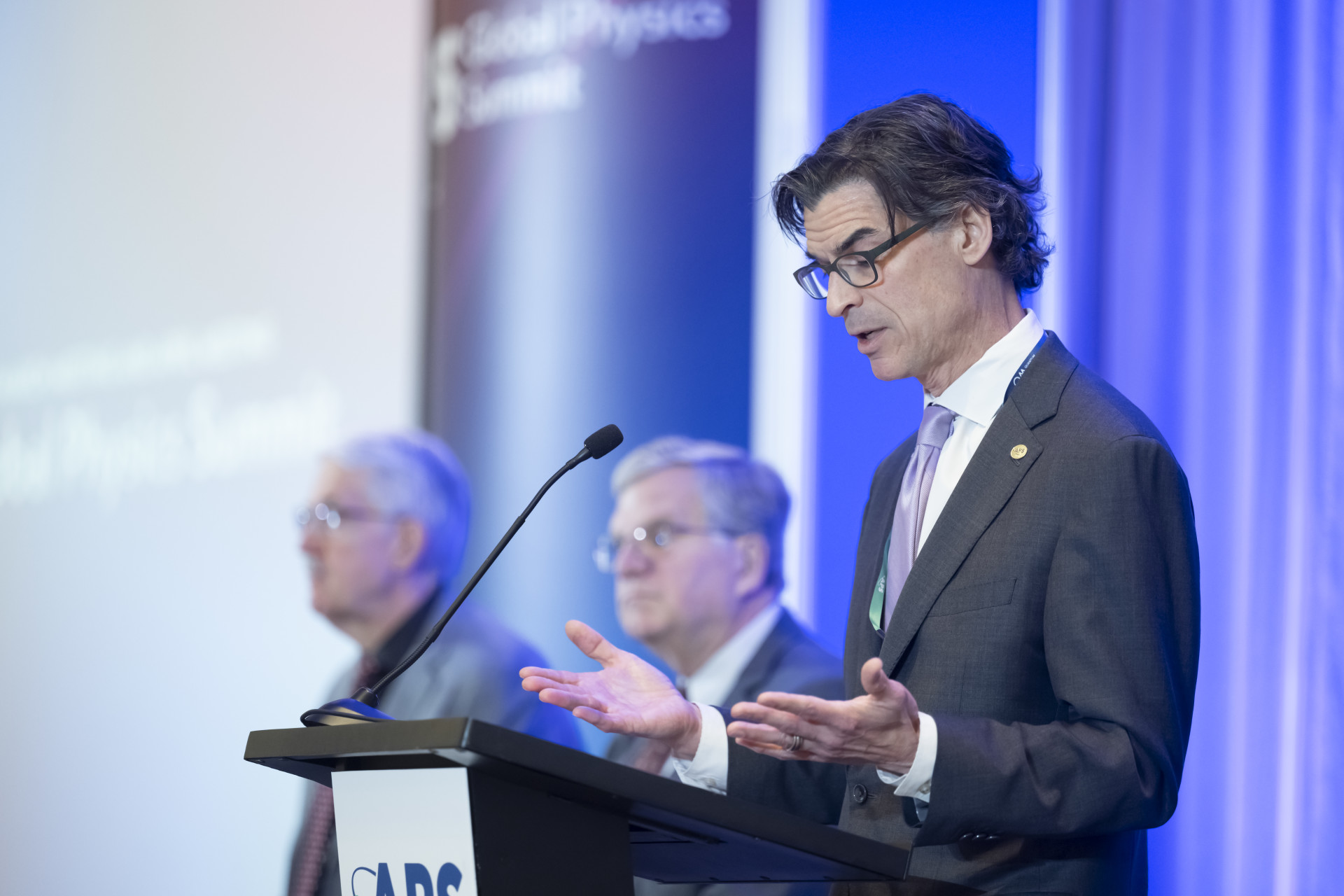 Francis Slakey speaks at a podium. John Doyle and Jon Bagger are seated in the background.