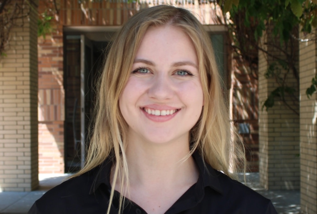 A headshot of Taylor Eskew, smiling with a brick building and foliage in the background.