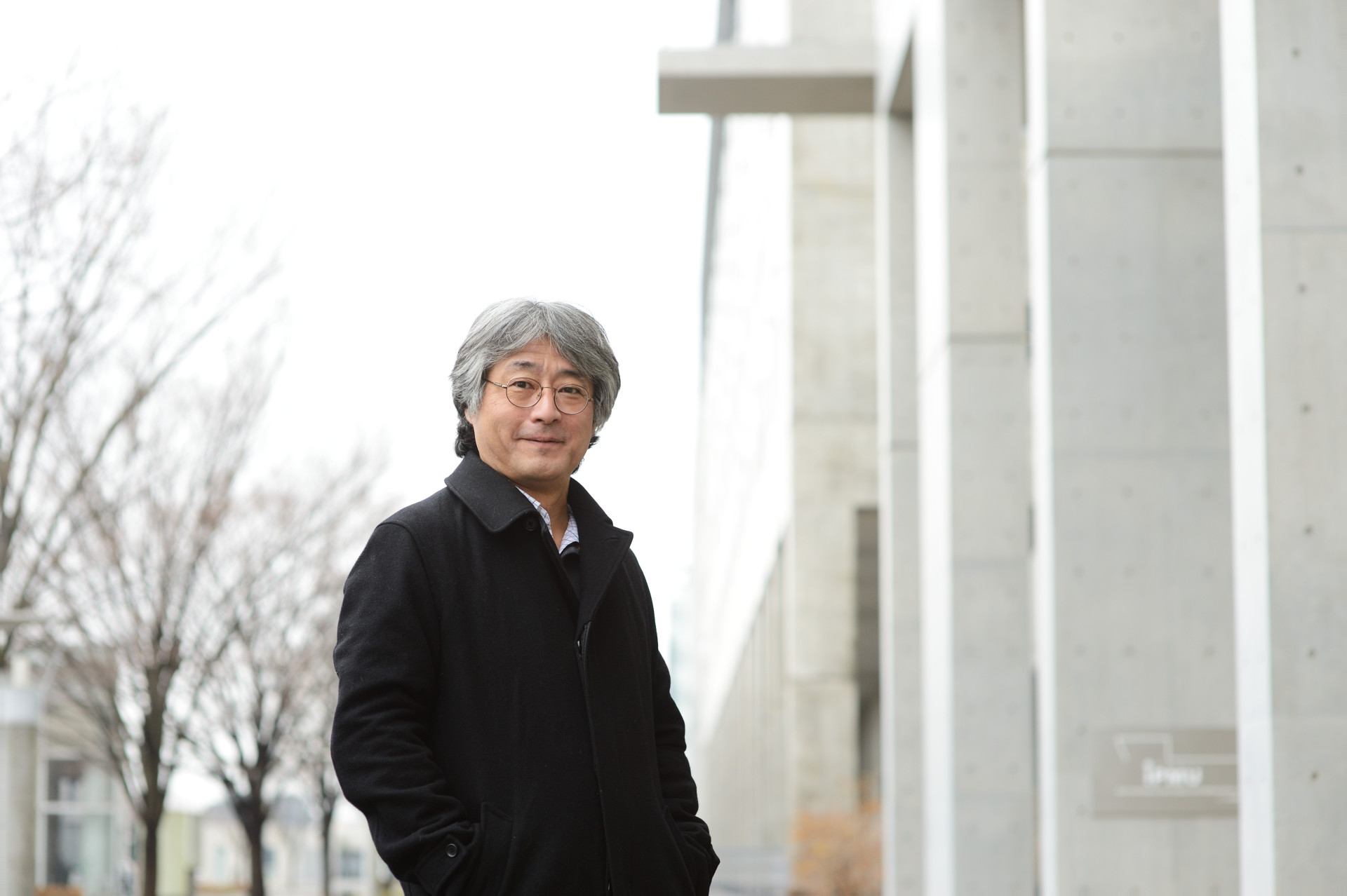 Murayama, wearing a black coat, stands outside on a gray day, with a concrete building in the background.