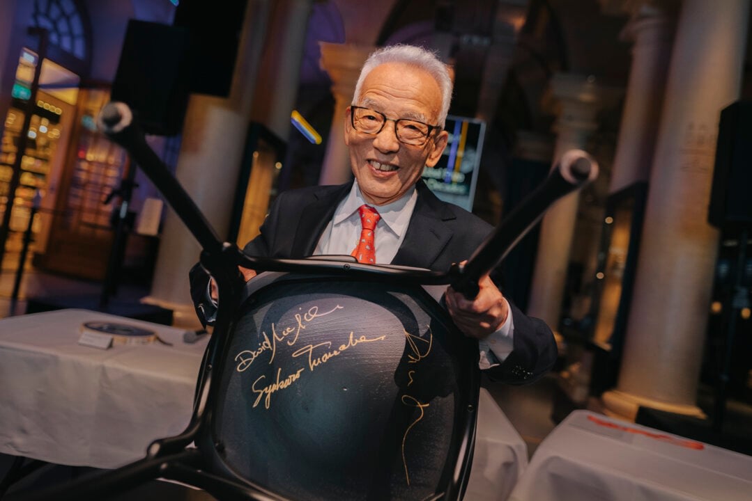 Manabe smiles and holds up a black chair, with his gold-colored autograph visible on the underside of the seat.