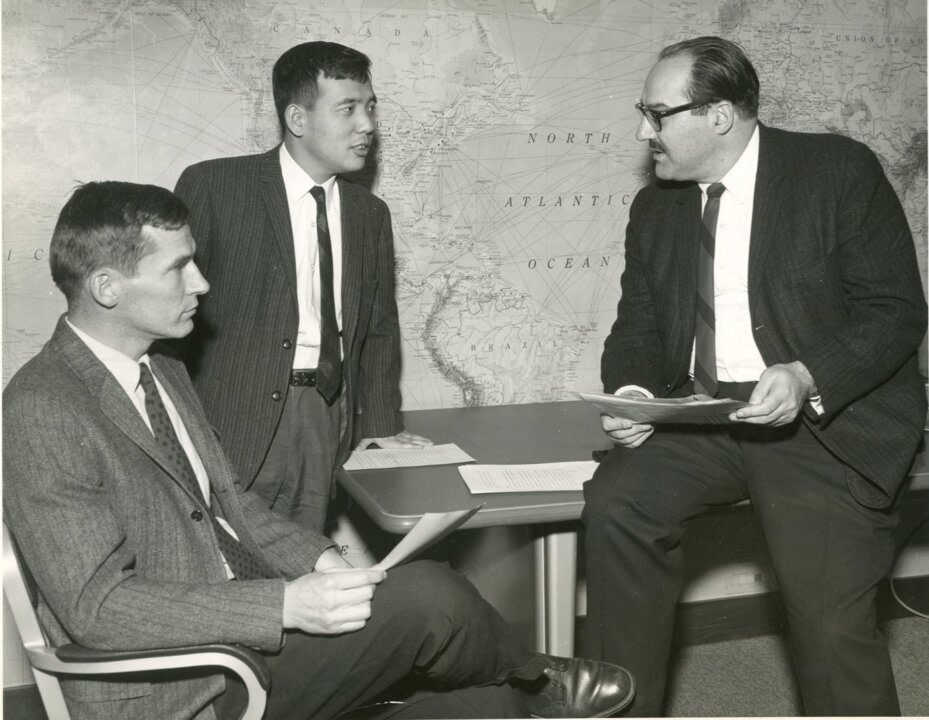 A black-and-white photo showing, from left to right, Bryan in a chair, Manabe leaning against a desk, and Smagorinsky sitting on the desk. The three, all in suits, are engrossed in conversation.
