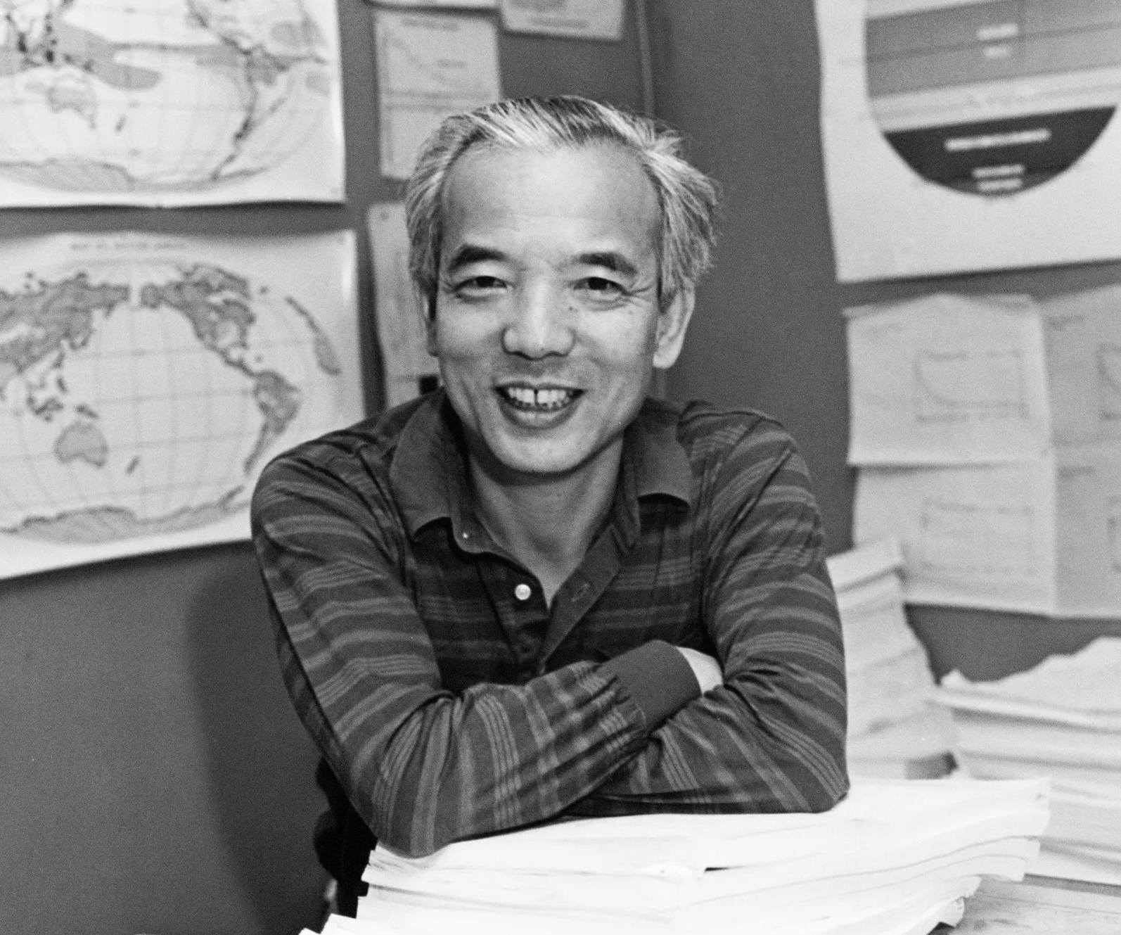 In this black-and-white photo, a younger Syukuro Manabe sits at a desk and smiles at the camera. His elbows are propped on a stack of papers, and maps and charts are visible on the wall behind him.