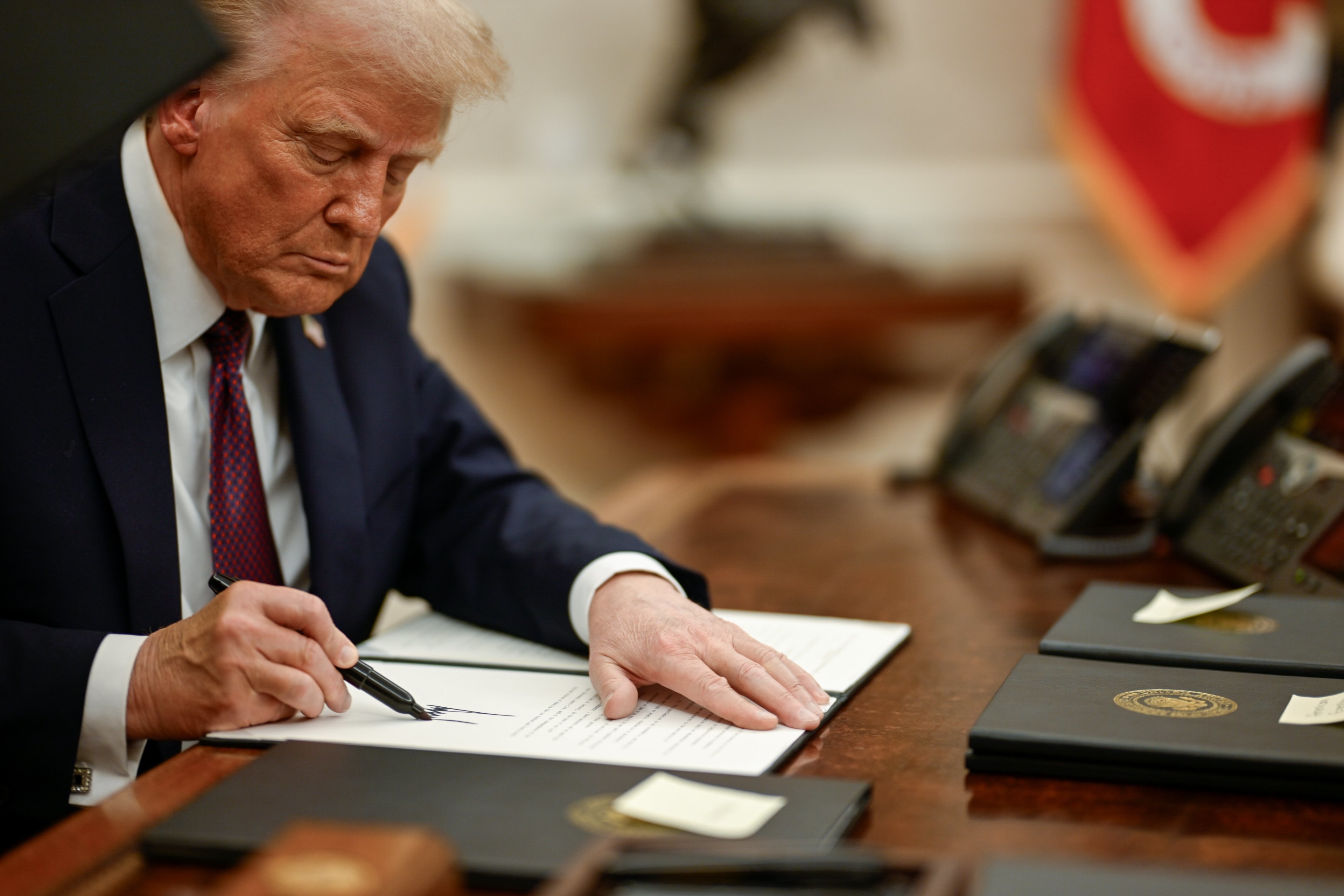 A close-up photograph of Trump, seated at a desk and signing a document.