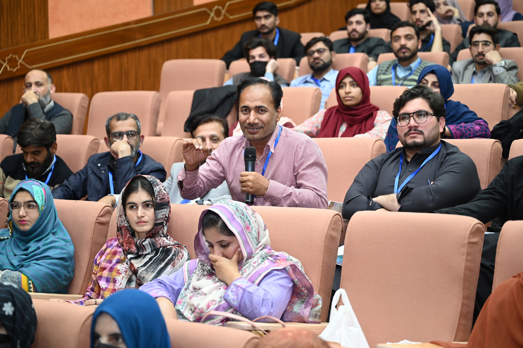 Audience member asking a question during a conference session, seated among attendees in an auditorium setting with many wearing conference badges.