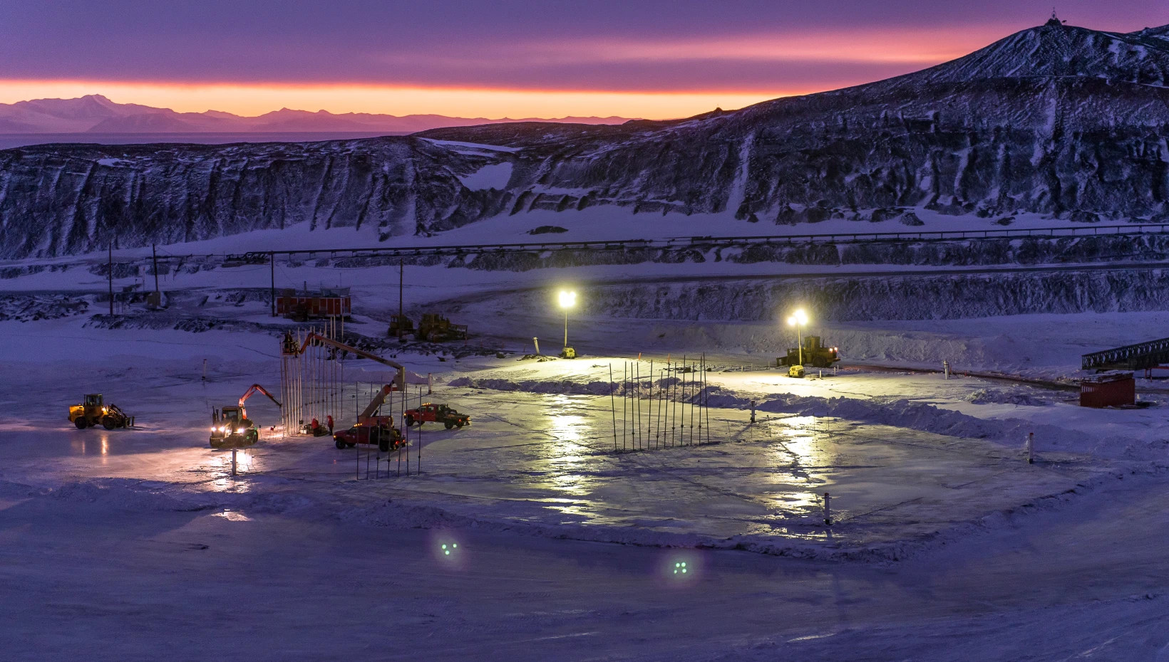Trucks and large machinery are parked on the icy ground at night under harsh spotlights.